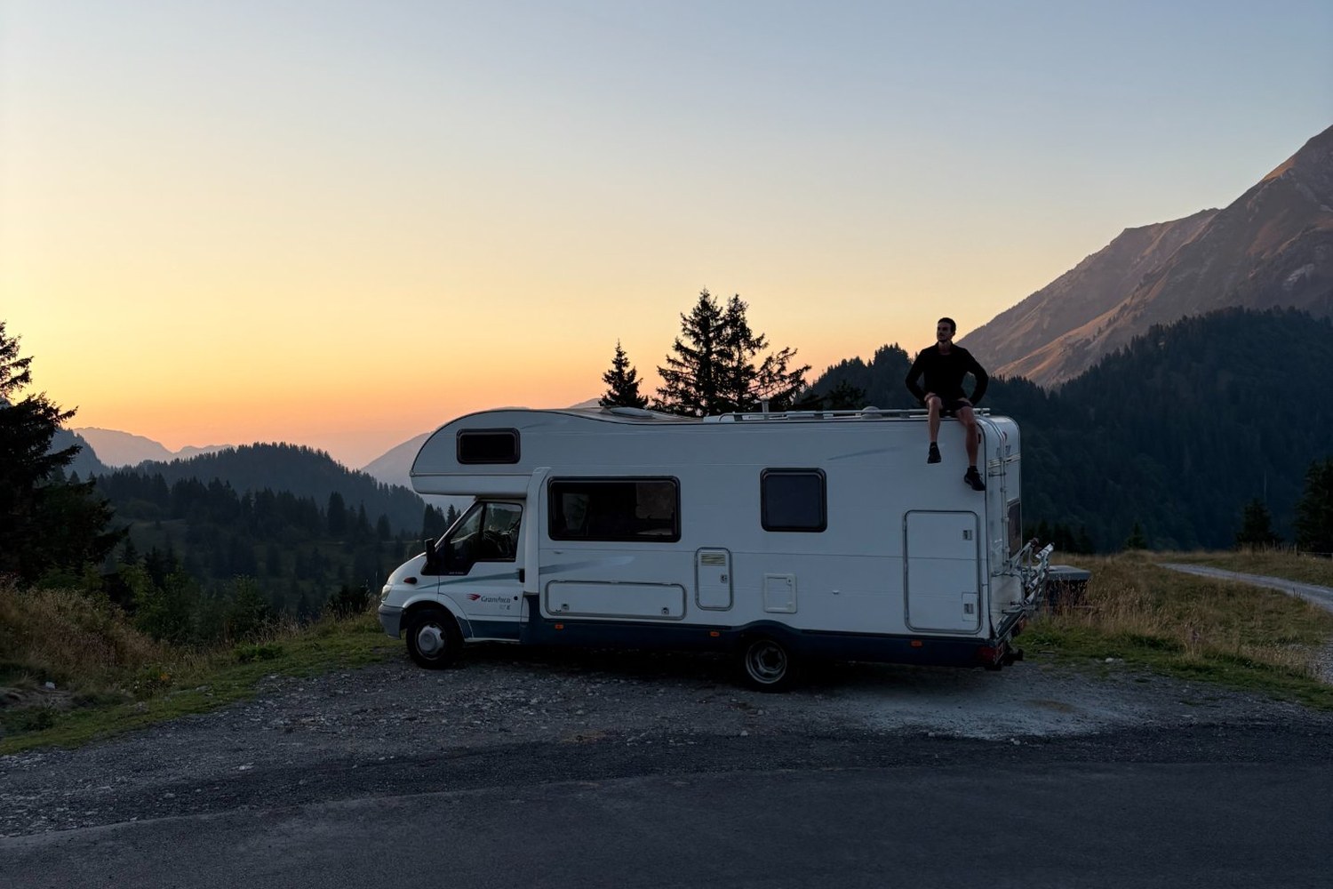The campervan parked on a mountain pass at sunset, someone sitting on the roof